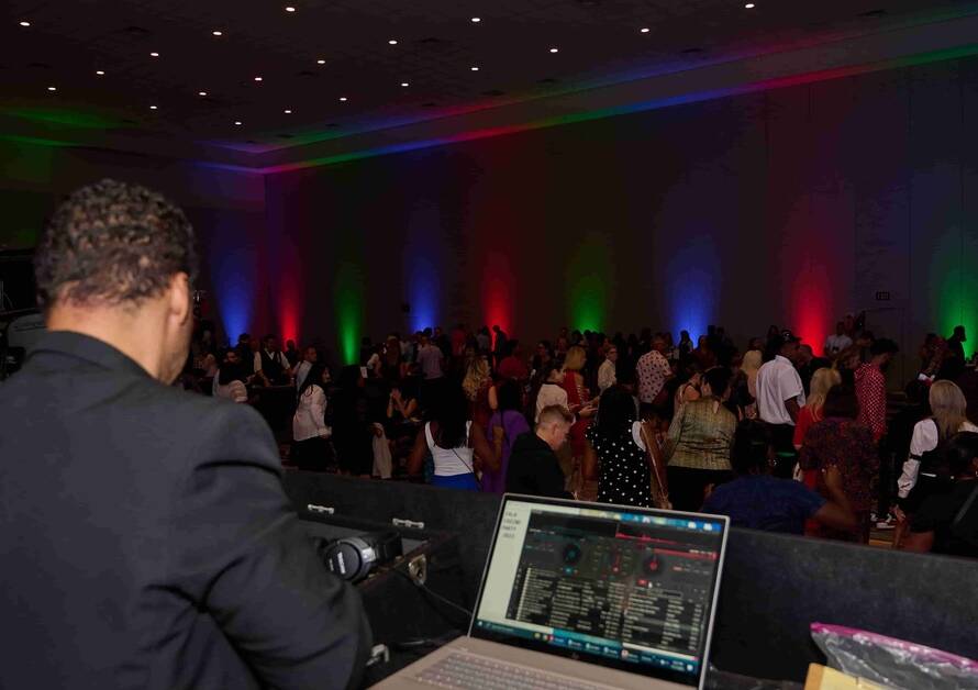 A DJ stands at a table in front of a crowd of people in a large ballroom. Red, green, and blue lights shine at the event.