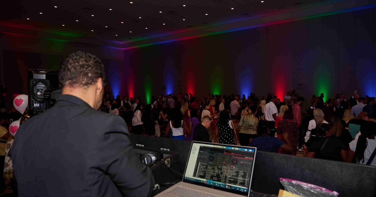 A DJ stands at a table in front of a crowd of people in a large ballroom. Red, green, and blue lights shine at the event.