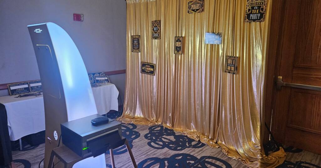A photo kiosk stands in front of a yellow curtain at an event. The curtain stands next to a wooden door.