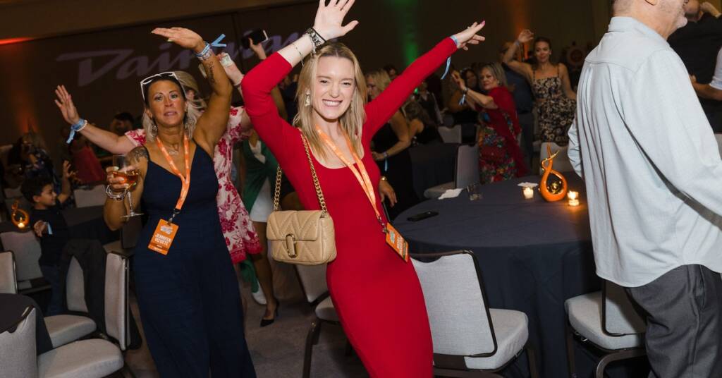 A group of people dance and look happy at an event with their hands in the air. They dance around several round tables.