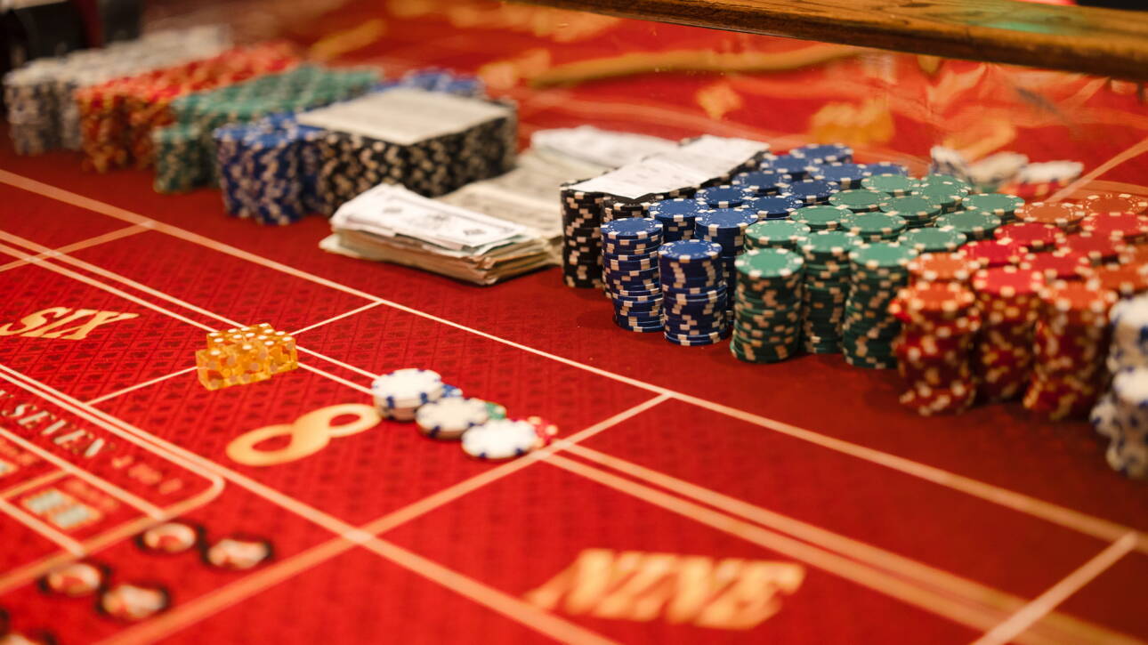 A casino table features stacks of chips of various colors, dice, and fake money. The table is red with yellow lettering.
