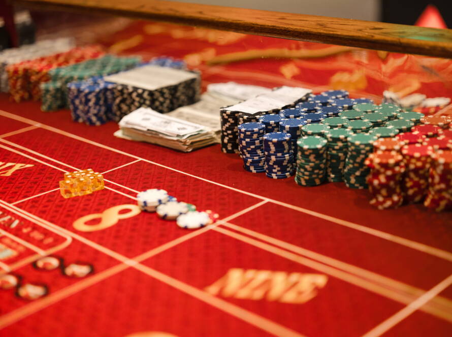A casino table features stacks of chips of various colors, dice, and fake money. The table is red with yellow lettering.
