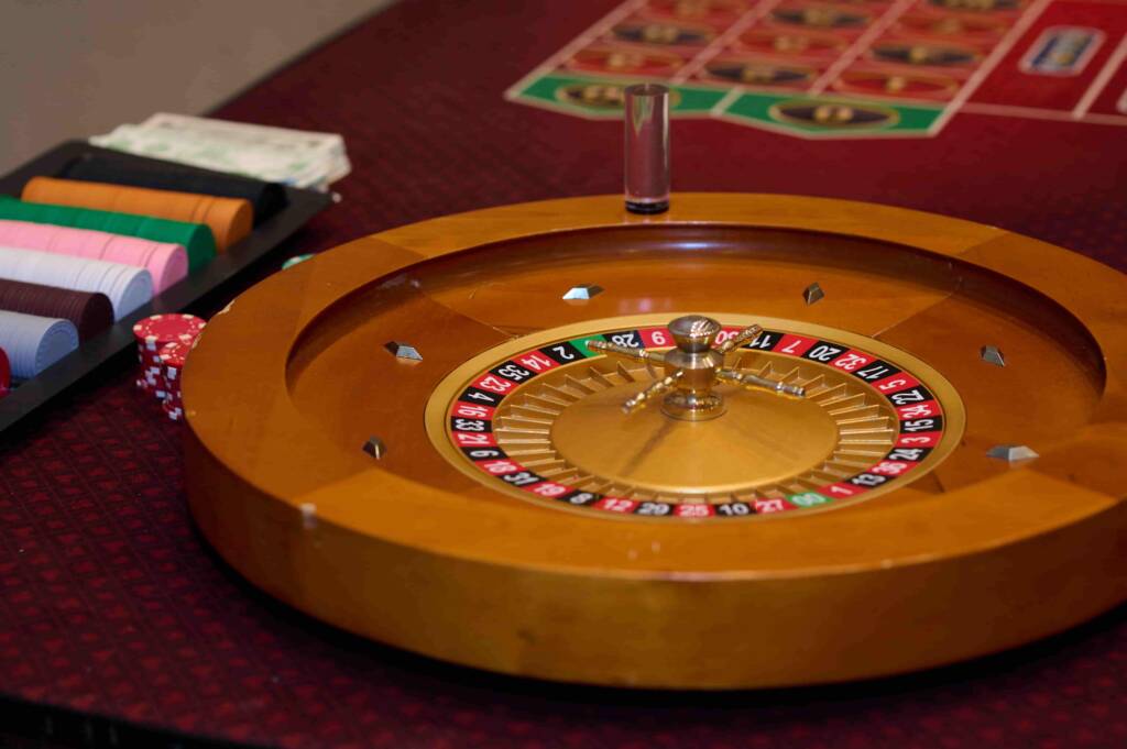 A wooden roulette wheel sits on a casino table next to stacks of chips and a grid with numbers. The table is red.