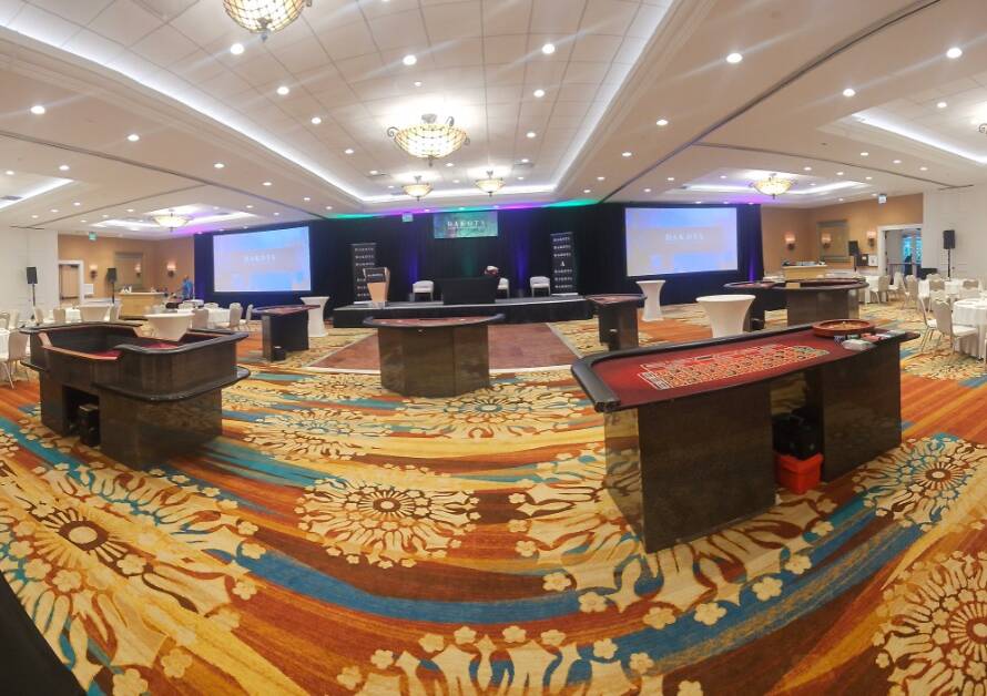 Casino tables fill a hotel ballroom as banquet seating, cocktail tables, and a stage surround the open event floor.