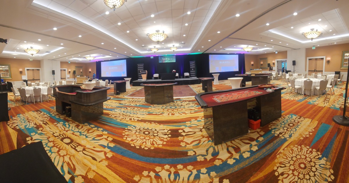 Casino tables fill a hotel ballroom as banquet seating, cocktail tables, and a stage surround the open event floor.