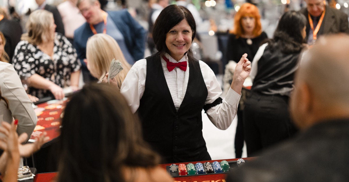 A smiling dealer stands behind a blackjack table as guests gather around the table at a casino night event in a ballroom.