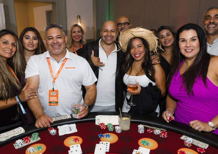 A group of smiling guests gathers around a casino-style table with chips, cards, and event badges in a ballroom.