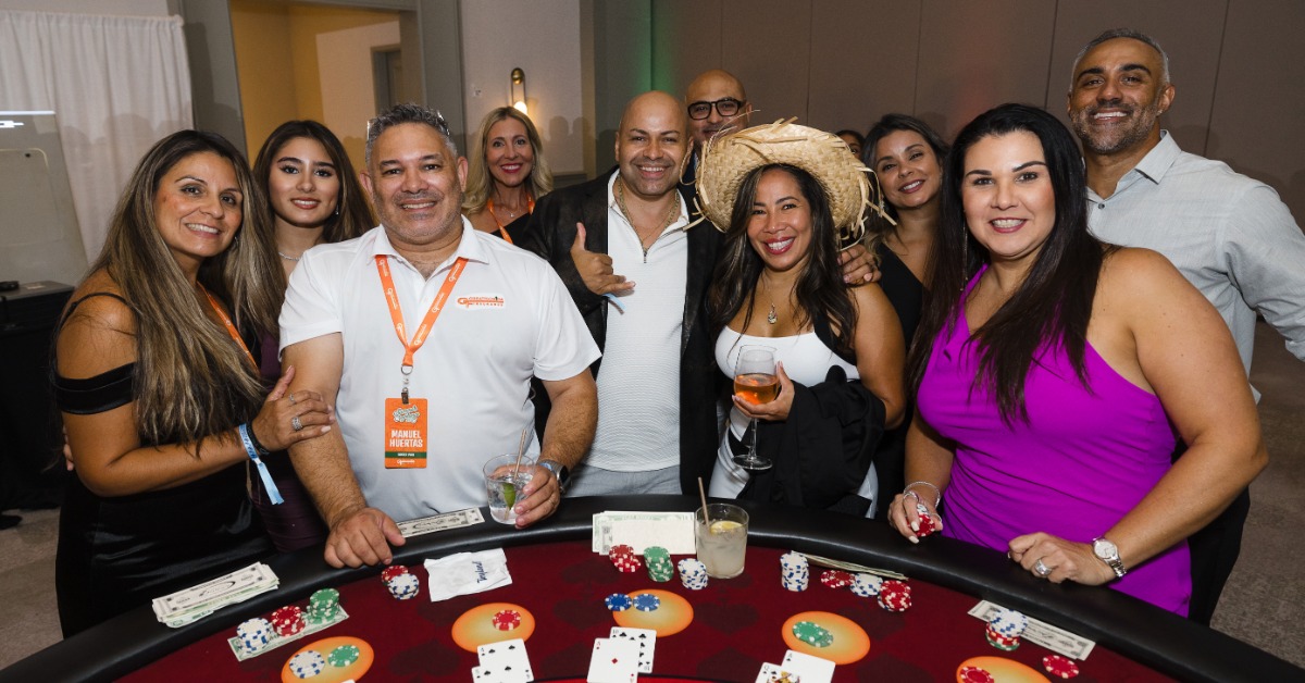 A group of smiling guests gathers around a casino-style table with chips, cards, and event badges in a ballroom.