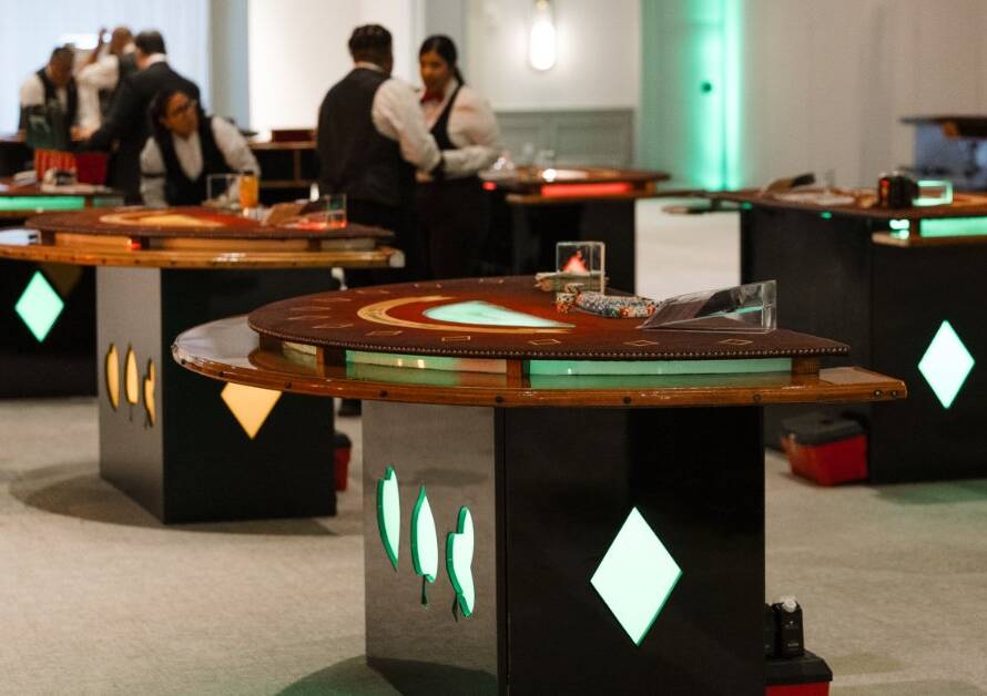 Casino tables fill an event space as professional dealers prepare the games under colorful accent lighting.