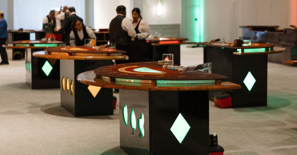 Casino tables fill an event space as professional dealers prepare the games under colorful accent lighting.