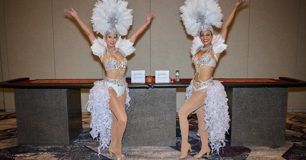 Two performers wearing white feathered costumes pose with raised arms in front of a large roulette table.