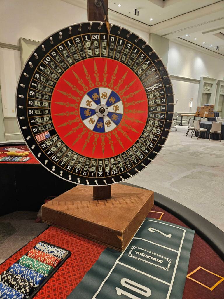 A large prize wheel sits on top of a blackjack table as colorful event chips and casino-game table markings are below.