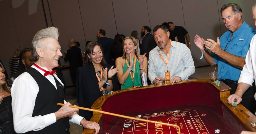 Casino party guests clap and cheer around a craps table as a dealer in a red bow tie leads the game.