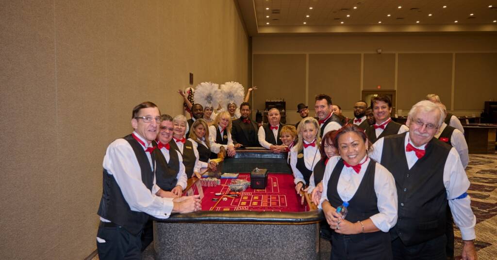 Casino event staff wearing black vests and red bow ties pose around a craps table, with showgirls in feathered costumes in the background.